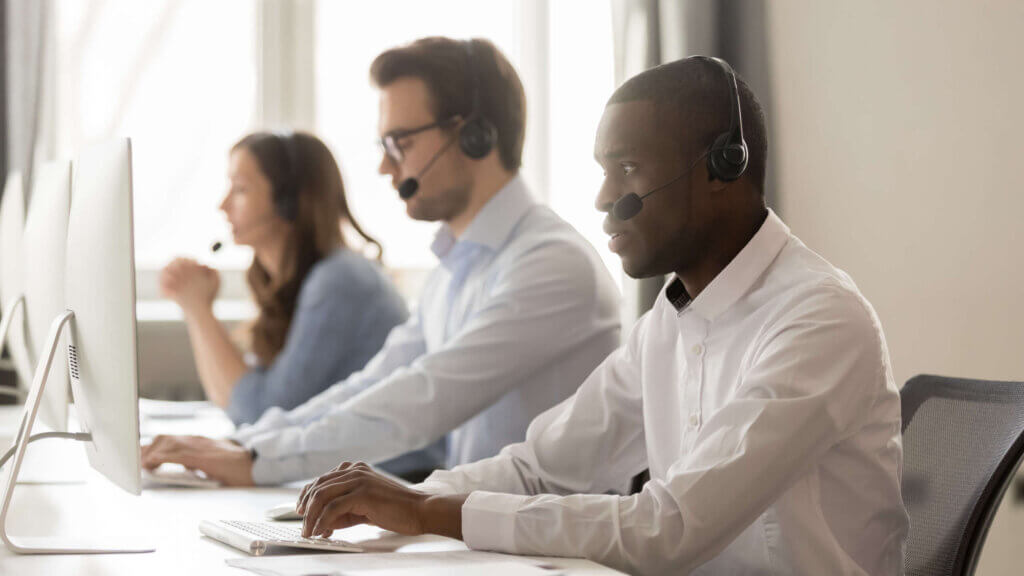Serious african call center agent in wireless headset working on computer with diverse team, focused black male operator telemarketer using pc in customer support service group office.